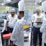 Team of chefs in white uniforms and hairnets inspect trays of food on a stainless steel prep line in a commercial kitchen