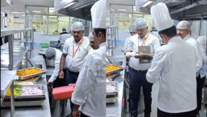 Team of chefs in white uniforms and hairnets inspect trays of food on a stainless steel prep line in a commercial kitchen