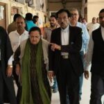 A group of formally dressed people walking down a hallway, led by a woman in green scarf, flanked by men in suits.
