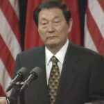 Older man in a dark suit and patterned tie, speaking at a podium with two microphones, flags in the background.