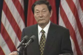 Older man in a dark suit and patterned tie, speaking at a podium with two microphones, flags in the background.