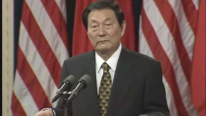 Older man in a dark suit and patterned tie, speaking at a podium with two microphones, flags in the background.